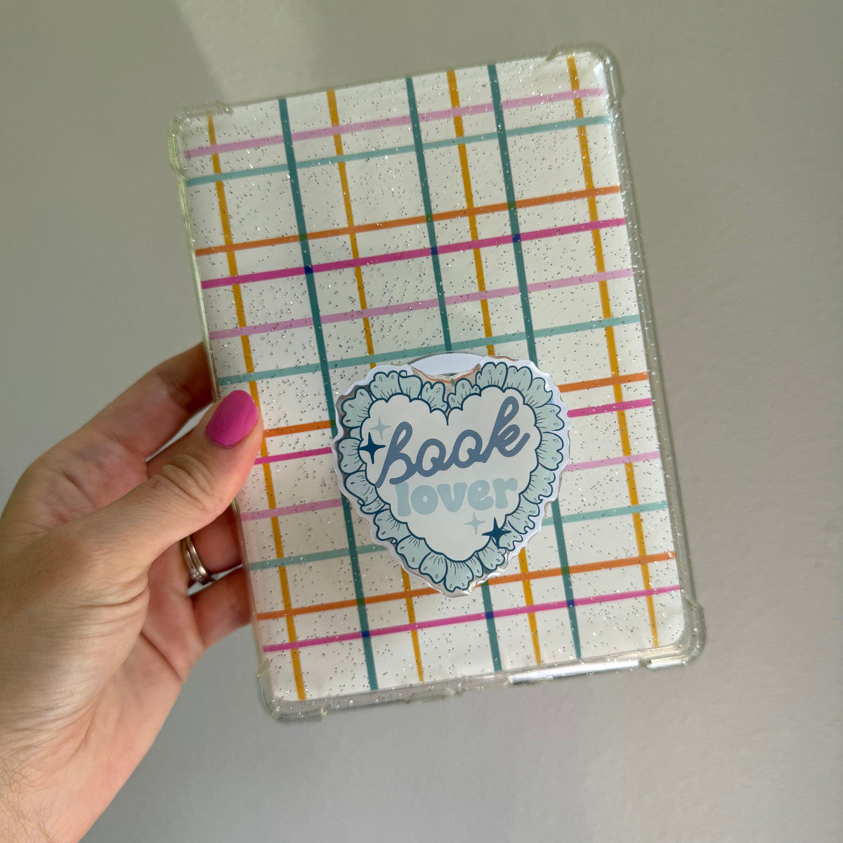 A hand with pink nail polish holds a clear protective case for a tablet. The case features a white background with a pastel multicolor plaid pattern and a central heart-shaped sticker that reads 'book over' in blue script.