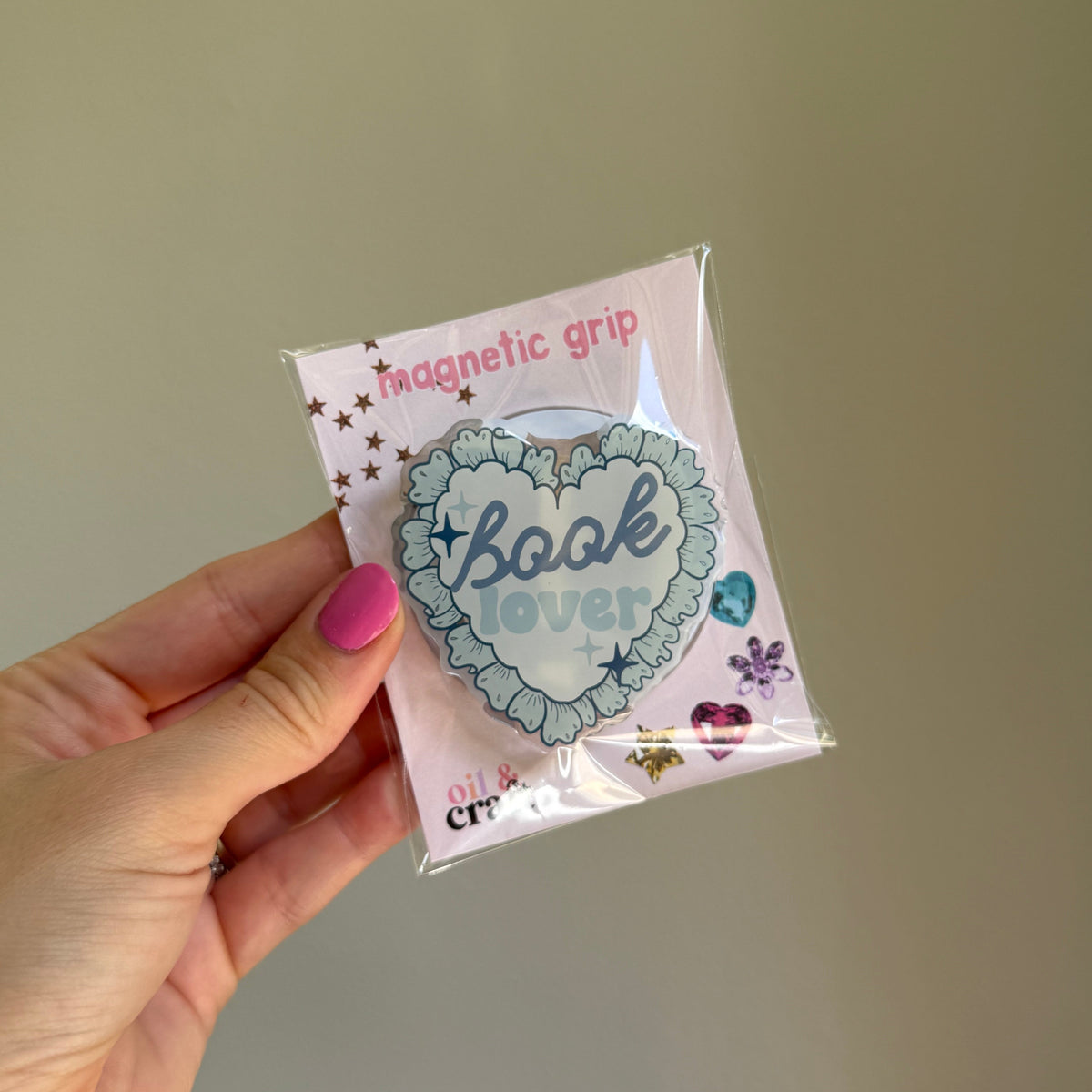 A hand with pink nail polish holds a packaged heart-shaped magnetic book grip. The grip features a light blue scalloped border and the words 'Book lover' in blue script, set against a pink backing card with small decorative illustrations.